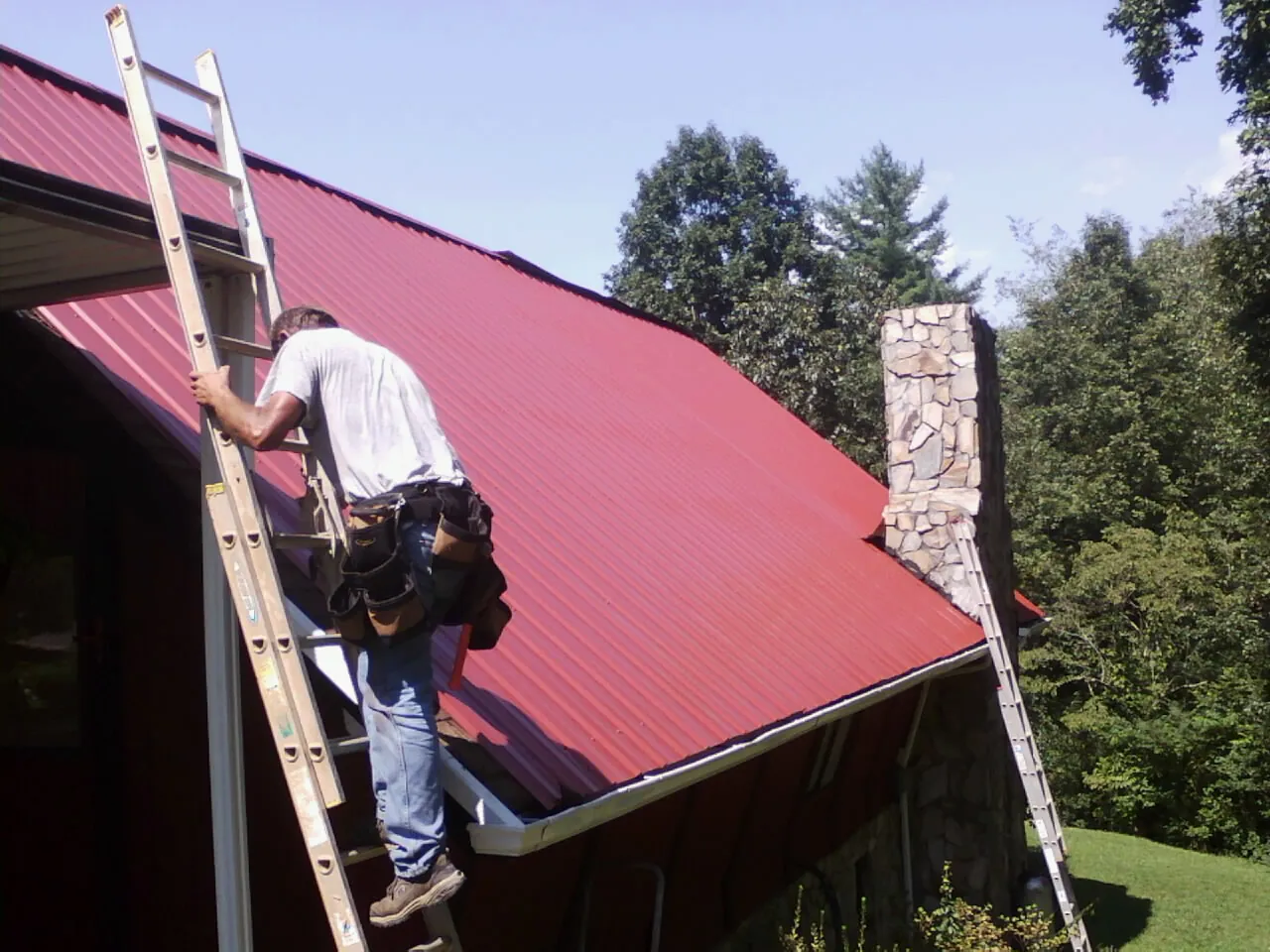 Roofer on a ladder beside a red metal roof and stone chimney — Soffit & Fascia Repair by Roofing Brothers in Stovall